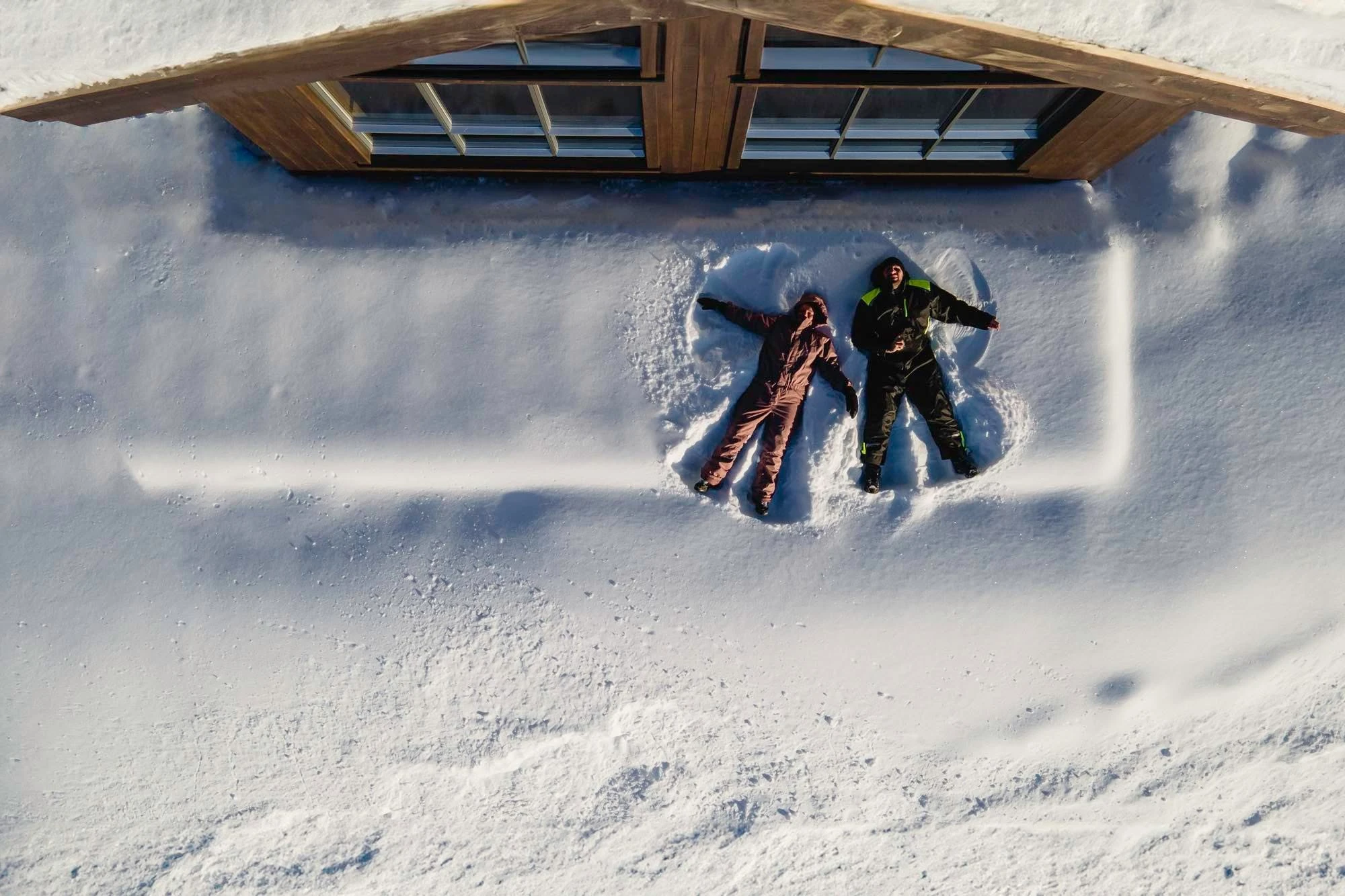 To personer lager snøengler i snøen utenfor hytta, sett ovenfra
